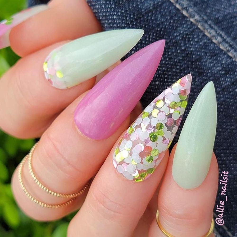 Nails with pink, green, and gold glitter design held by a hand against a denim background.