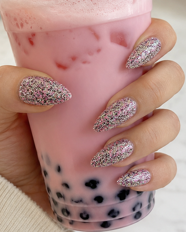 A hand with glittery pink and silver almond-shaped nail art holds a pink boba drink in a clear plastic cup. A small inset in the bottom right shows a close-up of the glitter nail design.
